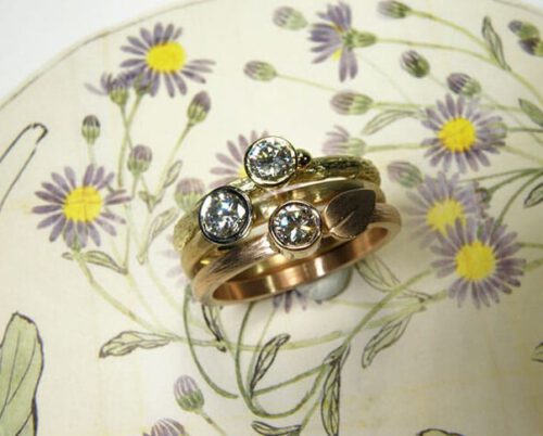 Verlovingsring 'Verzameling' geelgouden aanschuifring 'takje' met eigen diamant met besje, roodgouden aanschuifring 'ritme' met spitse hamerslag en met eigen diamant met blaadje, roségouden aanschuifring 'ritme' met kussen hamerslag en eigen diamant. Engagement ring ‘Collection’ yellow golden stack ring ‘twig’ with heirloom diamond and berry, rose golden stack ring ‘rhythm’ with hammering and heirloom diamond and leaf, rose golden stack ring ‘rhythm’ with cushion hammering and heirloom diamond. Oogst goudsmeden Amsterdam.