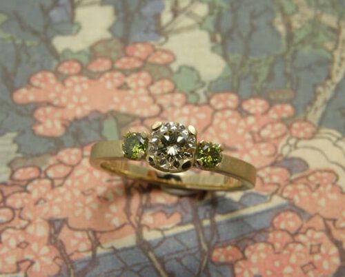 Verlovingsring 'Eenvoud'. Geelgouden ring met een 0,40 crt diamant en 2x 0,08 crt groene diamanten. Engagement ring 'Simplicity'. Yellow golden ring with a 0,40 crt diamond and 2x 0,08 crt diamonds. Uit het Oogst atelier Amsterdam.