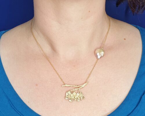 Gouden In bloei collier. Geelgouden bloesems op een takje aan een schakelcollier met een roze coin parel. Ontwerp van Oogst Sieraden in Amsterdam.